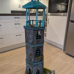 Puede incluir: Una maqueta detallada de una torre de piedra con un techo de estilo mirador turquesa. La torre presenta ventanas arqueadas, un efecto de piedra desgastada y vegetación artificial. La base es una formación rocosa con musgo.