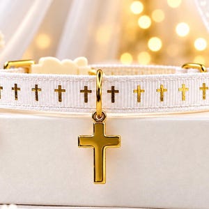 May include: A white pet collar featuring a repeating pattern of small gold crosses. The collar includes a gold-toned metal cross charm hanging from a ring and a small gold-toned bell attached to the side, with gold-toned hardware throughout.