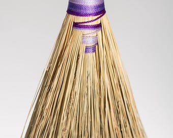 Può includere: Una scopa a mano realizzata con fibre di paglia color marrone chiaro naturale, con un manico avvolto da fili che alternano tonalità viola e lavanda. Un cordino intrecciato viola è fissato in cima per appendere il fascio di paglia.