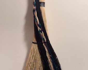 Può includere: Una scopa decorativa con setole di paglia color marrone naturale, arricchite da fibre nere e verde acqua. Il manico è avvolto in una corda nera e le setole sono intrecciate in modo elaborato con fili neri, verde acqua e viola.