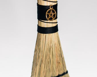 Può includere: Una piccola scopa decorativa in paglia naturale color marrone chiaro, con un cordino nero avvolto attorno al manico e alla base delle setole. Un ciondolo a forma di pentagramma in metallo dorato pende dal cordino sul manico.