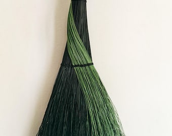Può includere: Una scopa decorativa con un manico lungo e sottile avvolto in corda nera, con una testa a spazzola ampia e svasata composta da setole sottili e rigide in sezioni alternate di nero profondo e verde bosco, legata con corda nera.