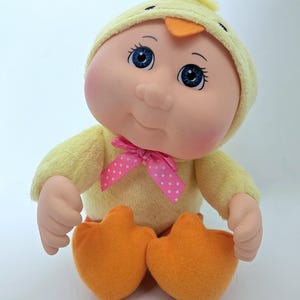 May include: A plush doll dressed as a baby chick. The doll has a yellow outfit, a matching hat with an orange beak, and orange feet. A pink bow with white polka dots adorns the doll's neck. The doll has blue eyes and rosy cheeks.