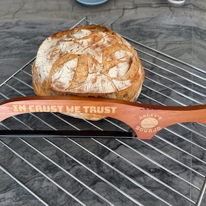 Könnte beinhalten: Ein Sauerteigbrot liegt auf einem Metall-Kühlrost, mit einem Brotmesser aus Holz im Vordergrund. Auf dem Brotmesser steht "IN CRUST WE TRUST" und "Haley's Sourjo".
