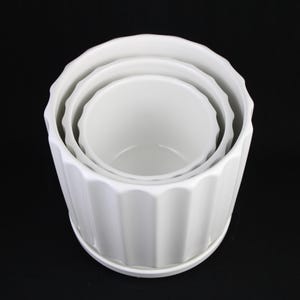 May include: A set of three white ceramic planters of varying sizes, nested together. Each planter has a ribbed exterior and a scalloped rim. The planters are sitting on a white saucer, all against a black background.