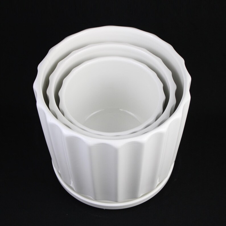 May include: A set of three white ceramic planters of varying sizes, nested together. Each planter has a ribbed exterior and a scalloped rim. The planters are sitting on a white saucer, all against a black background.