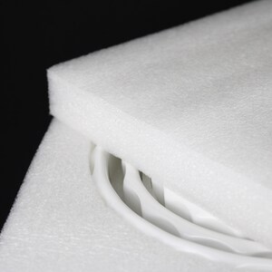 May include: Close-up of white foam packaging with curved white objects partially visible. The foam is thick and layered, providing protection. The background is black, highlighting the bright white of the foam and the objects.