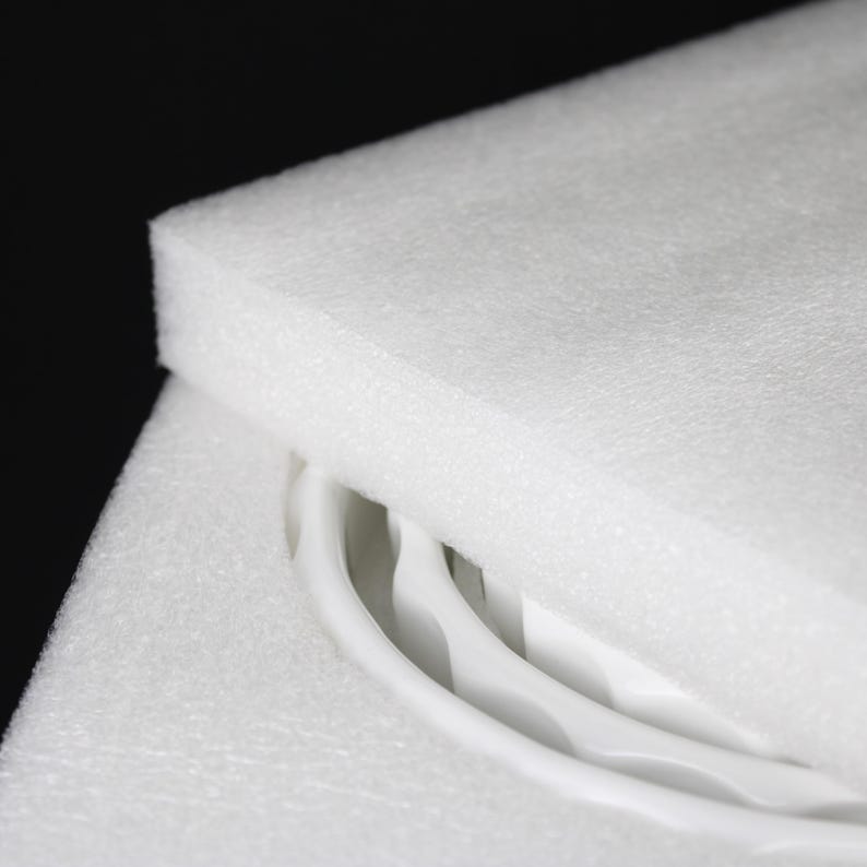May include: Close-up of white foam packaging with curved white objects partially visible. The foam is thick and layered, providing protection. The background is black, highlighting the bright white of the foam and the objects.