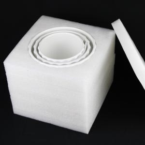 May include: A white foam cube with a circular cutout holding three white ceramic bowls. The bowls are nested within each other, with a wavy edge design. A separate foam piece rests beside the cube, against a black background.