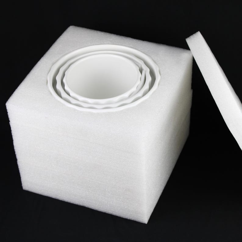 May include: A white foam cube with a circular cutout holding three white ceramic bowls. The bowls are nested within each other, with a wavy edge design. A separate foam piece rests beside the cube, against a black background.