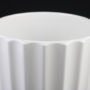 May include: A white ceramic vase with a fluted, geometric design. The vase has a wide opening and a smooth, glossy finish. The fluted sides create a play of light and shadow, adding visual interest to the simple, elegant form.