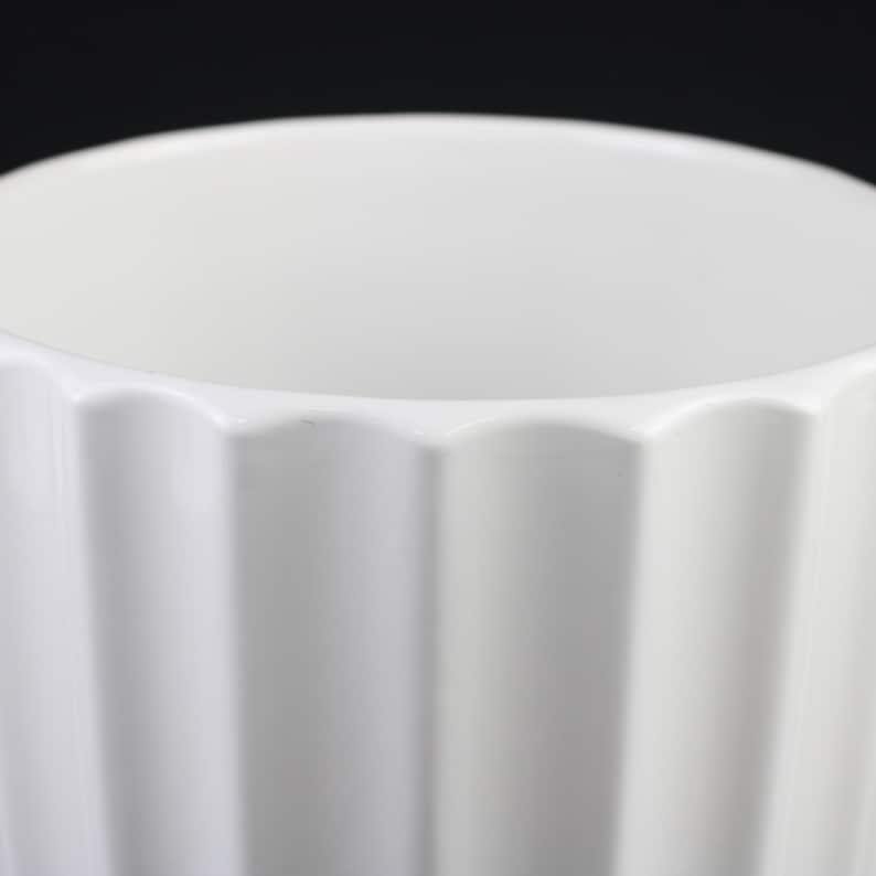 May include: A white ceramic vase with a fluted, geometric design. The vase has a wide opening and a smooth, glossy finish. The fluted sides create a play of light and shadow, adding visual interest to the simple, elegant form.