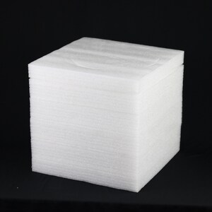 May include: A white foam cube, likely for packaging or crafting. The cube is composed of stacked layers of foam, creating a textured surface. The object is set against a black background, highlighting its shape and form.