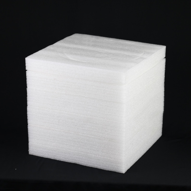 May include: A white foam cube, likely for packaging or crafting. The cube is composed of stacked layers of foam, creating a textured surface. The object is set against a black background, highlighting its shape and form.