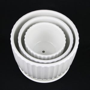 May include: Three white ceramic plant pots of varying sizes nested together. The pots have a ridged design around the exterior and a smooth interior. The largest pot has a matching saucer. The pots are empty and ready for planting.