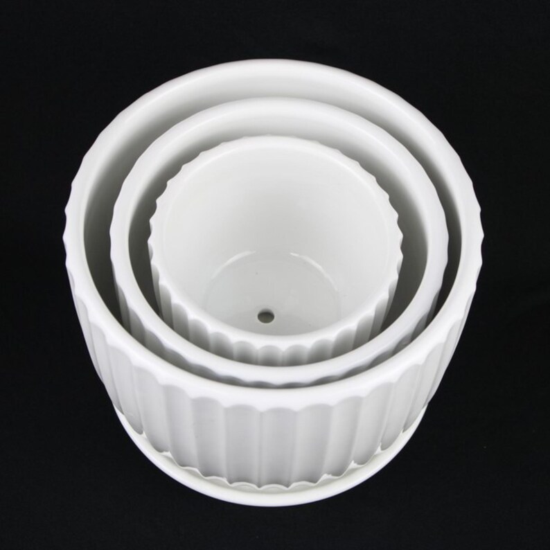 May include: Three white ceramic plant pots of varying sizes nested together. The pots have a ridged design around the exterior and a smooth interior. The largest pot has a matching saucer. The pots are empty and ready for planting.