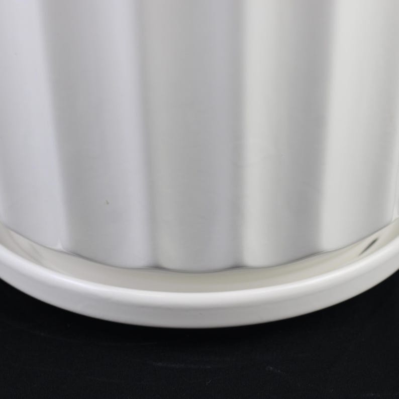 May include: A white ceramic planter with a ribbed design. The planter sits on a matching white saucer. The image is a close-up of the planter and saucer against a black background.