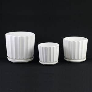 May include: Three white ceramic flower pots of varying sizes, each with a ridged exterior design and a matching saucer. The pots are cylindrical and have a clean, modern aesthetic, suitable for indoor or outdoor use.