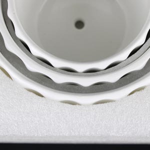 May include: Close-up of a white ceramic planter with a scalloped rim. The planter has a drainage hole at the bottom and is surrounded by protective gray foam. The design is simple and modern, suitable for various plants.