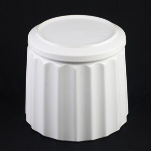 May include: A white ceramic container with a lid. The container has a fluted design on the body and a rounded lid. The object is set against a black background.