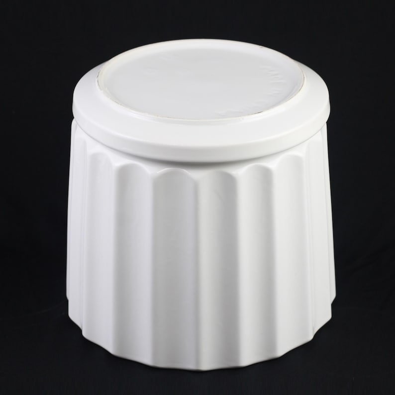 May include: A white ceramic container with a lid. The container has a fluted design on the body and a rounded lid. The object is set against a black background.