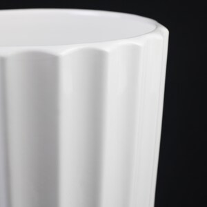 May include: A close-up of a white ceramic vase with a fluted design. The vase has a slightly scalloped rim and a glossy finish. The background is black, highlighting the vase's shape and texture.