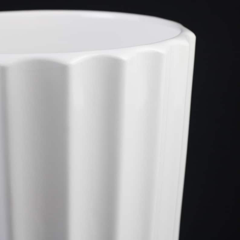 May include: A close-up of a white ceramic vase with a fluted design. The vase has a slightly scalloped rim and a glossy finish. The background is black, highlighting the vase's shape and texture.