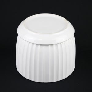May include: A white ceramic container with a lid. The container has vertical ribbed detailing and a rounded shape. The lid is a flat, circular shape. The background is black.