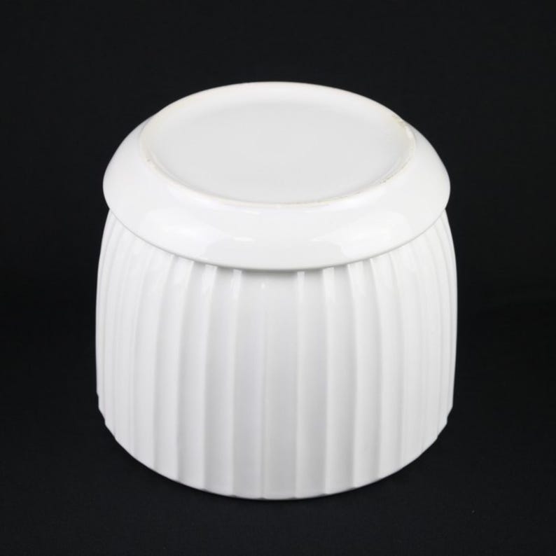 May include: A white ceramic container with a lid. The container has vertical ribbed detailing and a rounded shape. The lid is a flat, circular shape. The background is black.