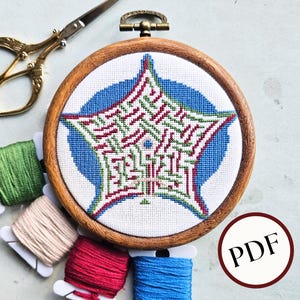 May include: A cross-stitch embroidery project in a wooden hoop, featuring a geometric star design in red, green, and white on a blue background. Includes gold scissors, colorful thread spools, and a PDF icon.