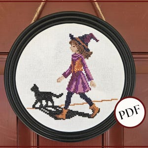 May include: Cross-stitch artwork in a black circular frame. The image depicts a young person in a purple witch costume walking with a black cat. The artwork is framed and ready to hang. A PDF icon is in the lower right corner.