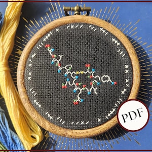 May include: A cross-stitch embroidery in a wooden hoop features a colorful molecular structure on black fabric. The design includes red, blue, and yellow accents. A PDF icon is visible. Embroidery floss in yellow, blue, and green is in the background.