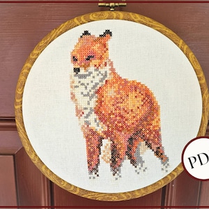Op de afbeelding: Kruissteekborduurwerk van een vos in tinten oranje, bruin en grijs, gezet in een houten hoepel. De vos is in een zijaanzicht afgebeeld, met gedetailleerde steken. De hoepel hangt aan een houten deur.