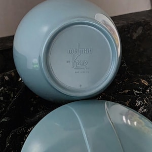 May include: Two light blue, round bowls. The bottom of one bowl is visible, displaying the embossed text "melmac by Kenro" and patent information. The bowls have a glossy finish and a simple, classic design. The bowls are likely vintage.