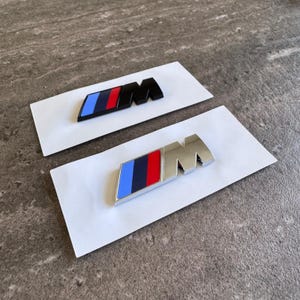 Peut inclure: Deux emblèmes M, l'un avec une finition noire mate et l'autre avec une finition chromée polie, chacun présentant un parallélogramme incliné avec trois bandes verticales bleu clair, bleu foncé et rouge, suivies de la lettre M sur fond blanc.