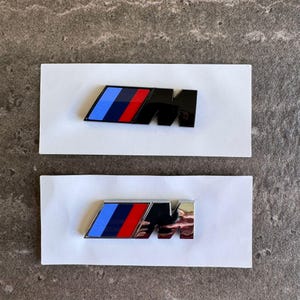 Pode incluir: Duas insígnias de emblema de carro com o icónico logótipo M da BMW. A insígnia superior é preta com riscas azuis, azul escuro e vermelhas. A insígnia inferior é cromada com o mesmo design de riscas. Ambas estão sobre um suporte branco.