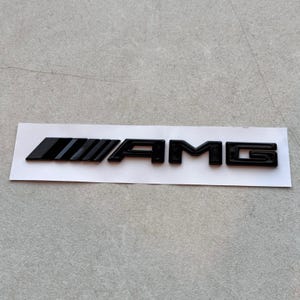 Puede incluir: Un emblema AMG negro brillante que presenta cuatro barras paralelas inclinadas seguidas de las letras A, M y G en una tipografía de bloque en negrita. El emblema está montado sobre una tira adhesiva blanca rectangular con un acabado liso y brillante.