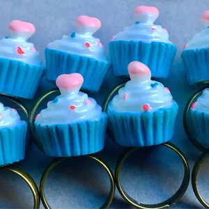 Könnte beinhalten: Neun blaue Cupcake-Ringe mit weißem Frosting und rosa und blauen Streuseln. Die Ringe sind aus Metall und haben eine goldene Oberfläche.
