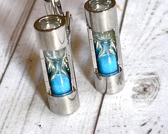 May include: A pair of silver cufflinks with a small hourglass design. The hourglass is filled with blue sand.