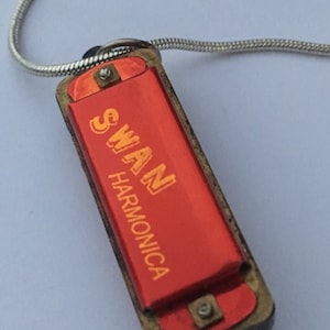 May include: A red harmonica with a gold-colored metal frame and a silver chain. The harmonica has the word "SWAN HARMONICA" printed on it in orange letters.