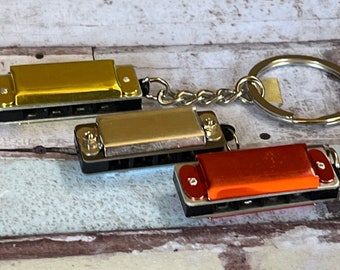 May include: Miniature harmonica keychains in gold, silver, and orange. Each keychain features a small, detailed harmonica attached to a chain and keyring. The harmonicas have black accents and are displayed against a light wood-grain background.