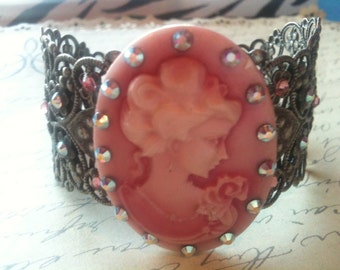 May include: A vintage-style cuff bracelet featuring a pink cameo of a woman's profile, surrounded by iridescent rhinestones. The ornate, filigree metalwork is a dark, antique silver color, creating a romantic and elegant accessory.