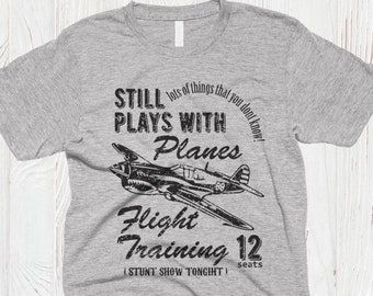 May include: A heather grey short-sleeved t-shirt featuring a black graphic of a vintage fighter plane. The text reads: STILL lots of things that you dont know! PLAYS WITH Planes FLIGHT TRAINING 12 seats (STUNT SHOW TONIGHT).