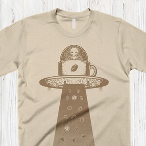 May include: A beige t-shirt with a graphic of a UFO shaped like a coffee cup with an alien inside. Coffee beans are falling from the UFO.
