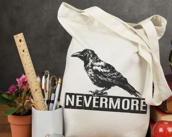 Może przedstawiać: Biała torba z płótna z czarnym rysunkiem kruka i napisem "NEVERMORE". Torba leży na stole z książkami, jabłkiem i ołówkiem.