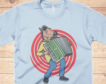 Op de afbeelding: Lichtblauw T-shirt met een afbeelding van een paard dat een geel shirt draagt en accordeon speelt. Het paard staat voor een rood-witte spiraalvorm.