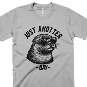 May include: A gray t-shirt with a black and white illustration of an otter wearing sunglasses. The text "Just Anotter Day" is printed on the shirt.