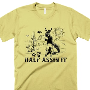 May include: A yellow t-shirt with a black graphic of a donkey sitting in a desert landscape with cacti and the text "HALF-ASSIN IT".