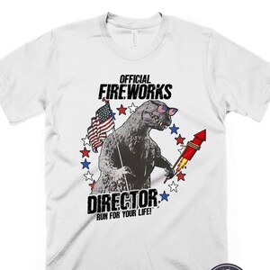 Puede incluir: Camiseta blanca con un gr&aacute;fico de un monstruo parecido a Godzilla que lleva gafas de sol y sostiene un fuego artificial. El gr&aacute;fico est&aacute; rodeado de estrellas rojas, blancas y azules. El texto "OFFICIAL FIREWORKS DIRECTOR RUN FOR YOUR LIFE!" est&aacute; impreso debajo del gr&aacute;fico.