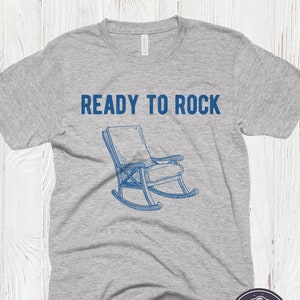 May include: A gray t-shirt with the text "READY TO ROCK" in blue lettering. A blue illustration of a rocking chair is below the text. The t-shirt has a distressed look.